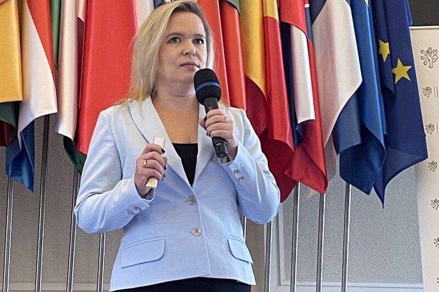 Adriana Jahňáková speaking with a microphone in front of a row of European national flags.